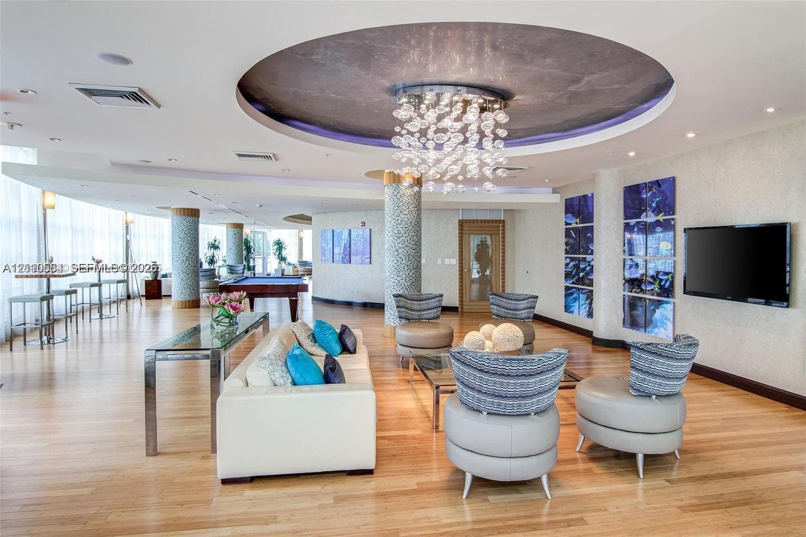Expansive room featuring wood-finish flooring, a recessed ceiling with a circular feature, a multi-tiered crystal chandelier, decorative tile-clad structural columns, and wall-to-wall floor-to-ceiling windows