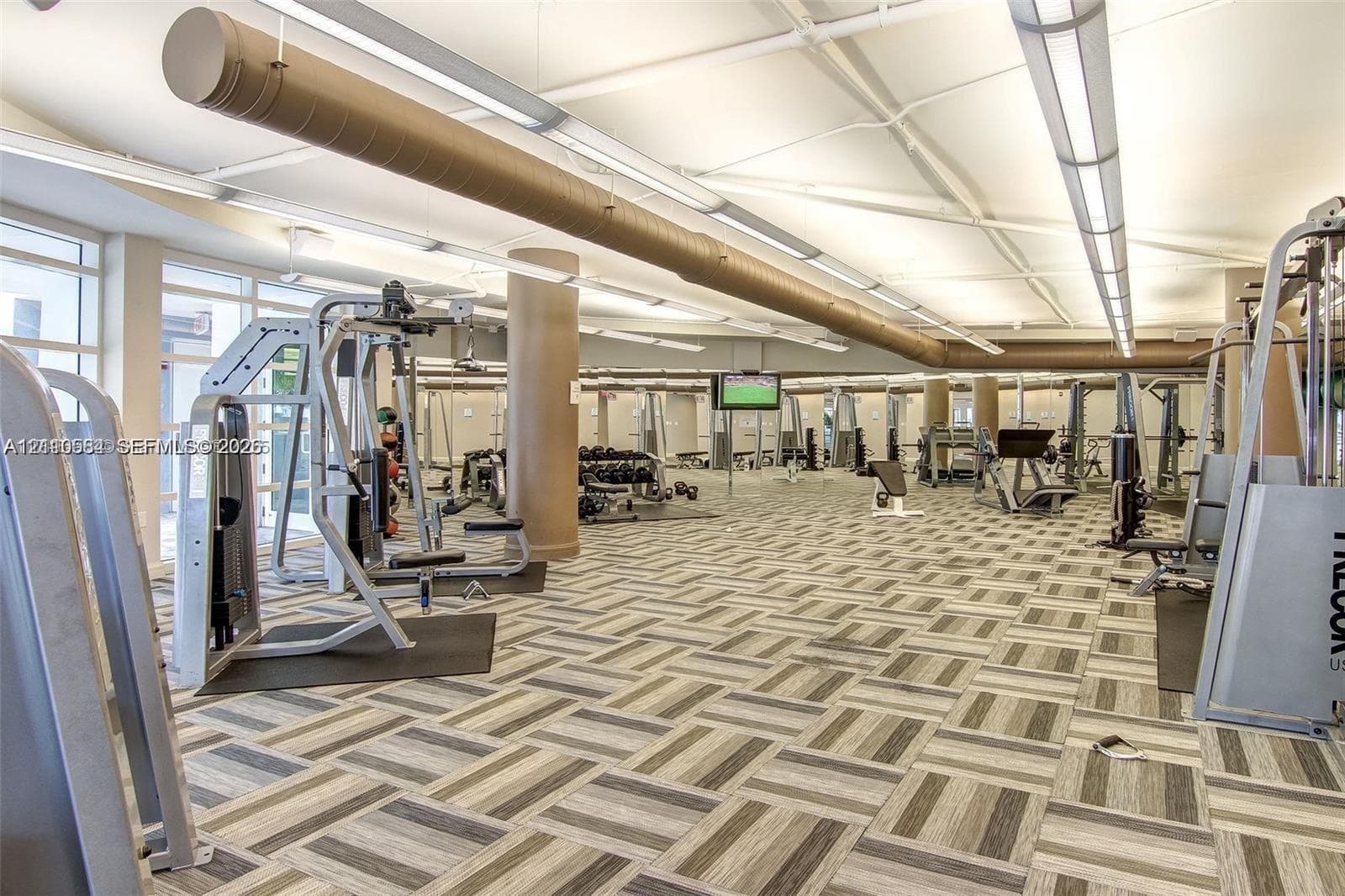 Fitness center featuring large-format linear lighting, exposed architectural ductwork, patterned industrial carpet tiles, and structural support columns