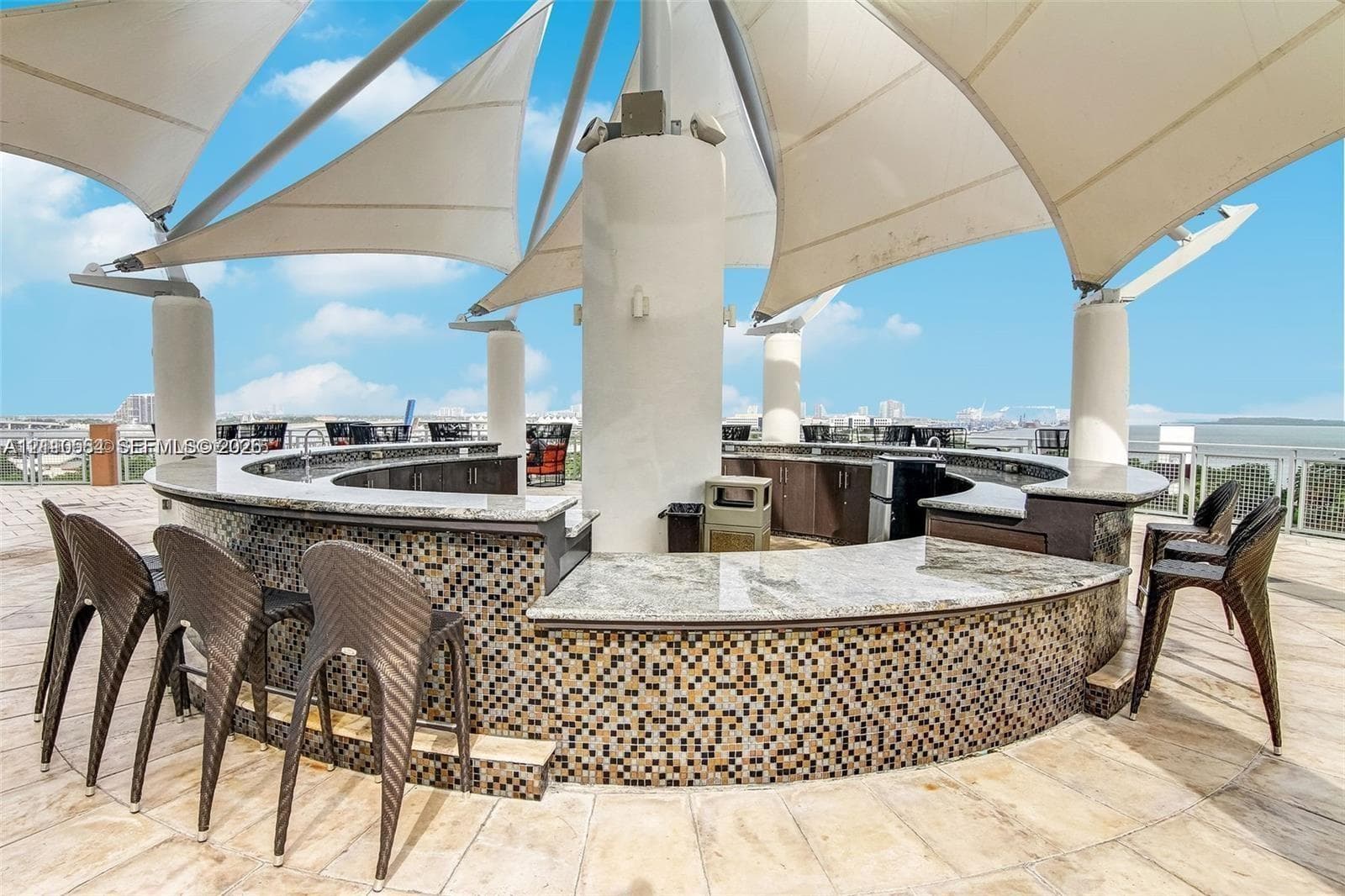Rooftop bar area featuring a curved counter with stone-surface top, decorative mosaic tile base, tension-fabric shade structure, and stone tile flooring