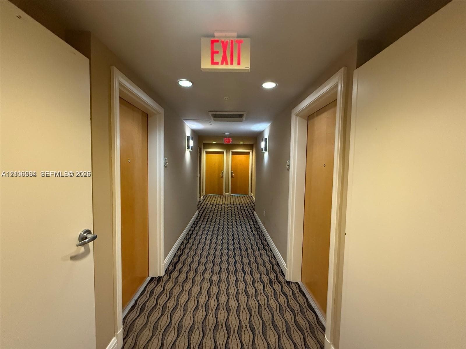 Hallway featuring patterned wall-to-wall carpeting, wood-finish interior doors with metallic hardware, wall-mounted light fixtures, and ceiling-mounted recessed lighting