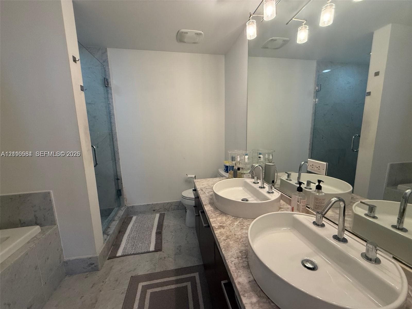 Double vanity featuring oval vessel sinks, stone-finish countertops, dark cabinetry, and a full-width wall mirror, complemented by a glass-enclosed shower with tiled surround