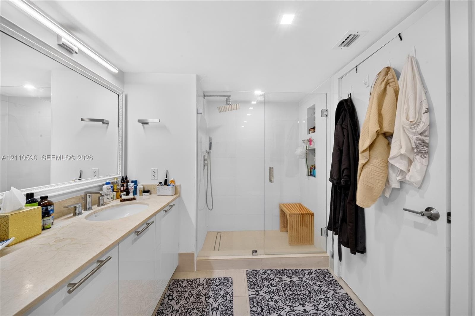 Full-width vanity with an integrated sink and light-toned solid surface countertop, walk-in glass-enclosed shower featuring a rainfall showerhead, and recessed tiled shelving