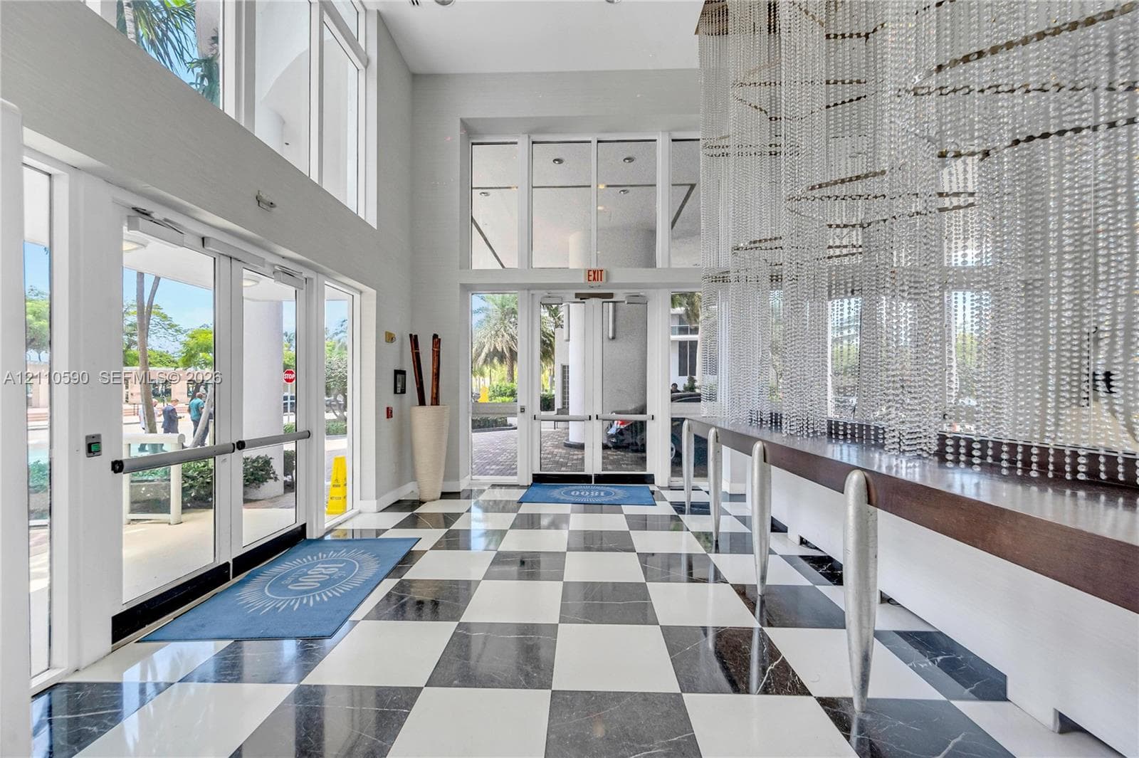 Grand entrance featuring double-height ceilings, floor-to-ceiling glass paneling, high-contrast checkerboard stone flooring, and a large-scale crystal chandelier