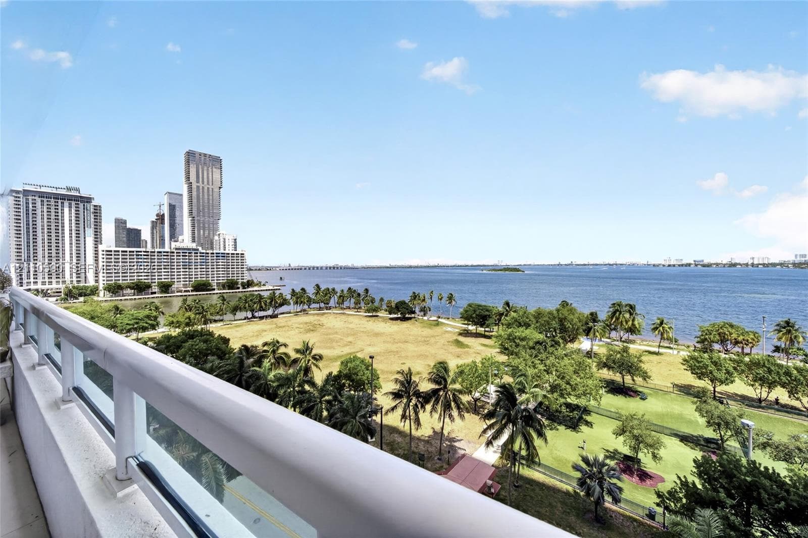 Balcony featuring a white metal railing with glass panels, overlooking a large waterfront park, mature palm trees, and an urban skyline