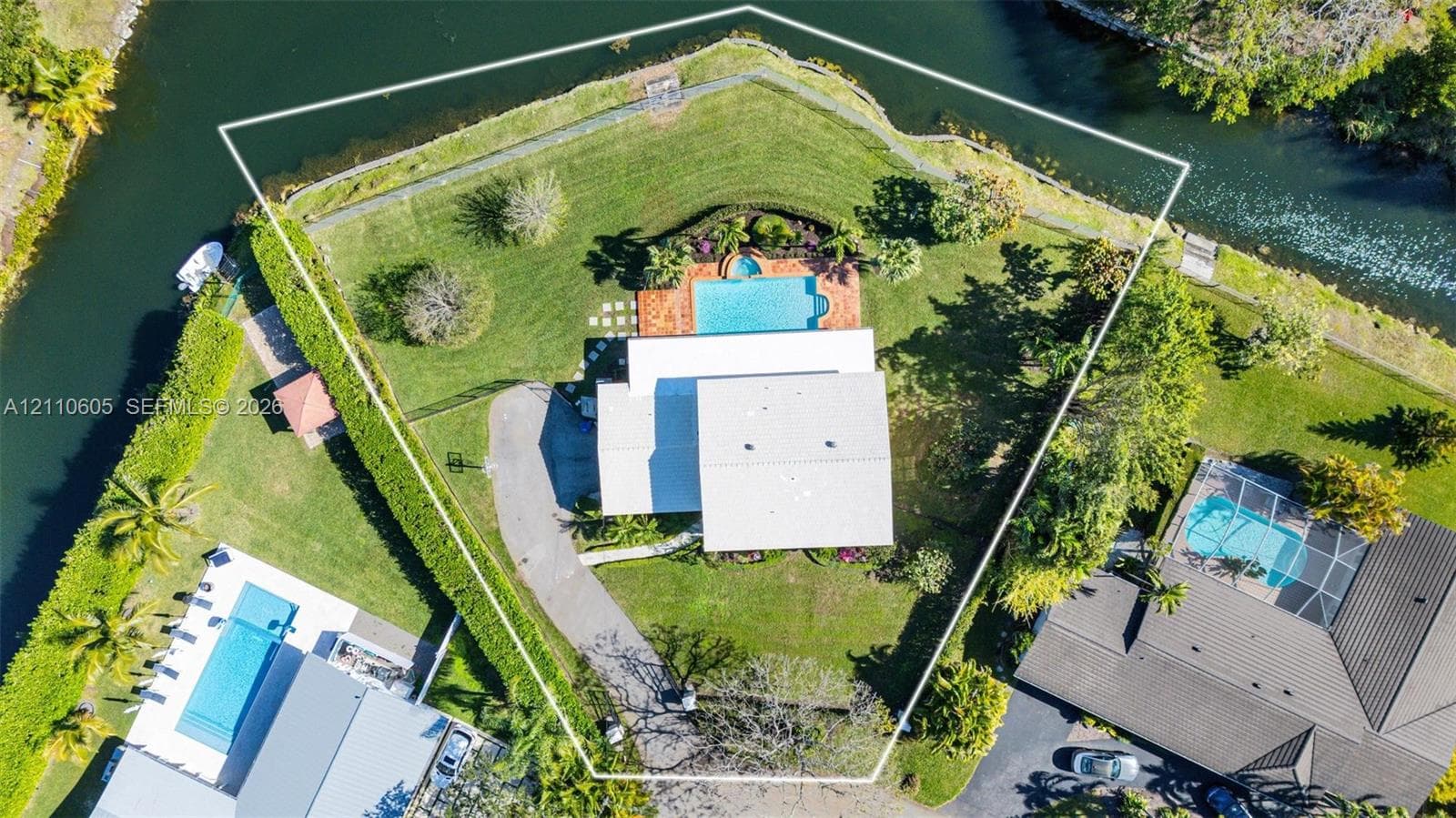 Waterfront lot featuring a perimeter fence, expansive lawn, mature landscaping, and a paved driveway leading to a swimming pool