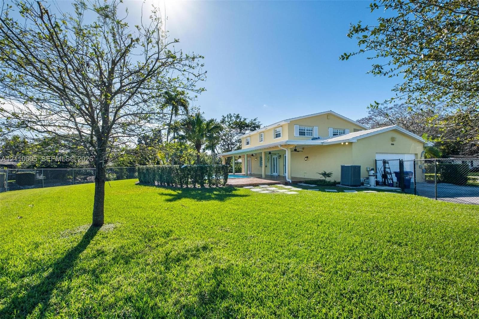 Spacious backyard featuring a manicured lawn, perimeter chain-link fencing, mature trees, a covered patio, and a concrete driveway
