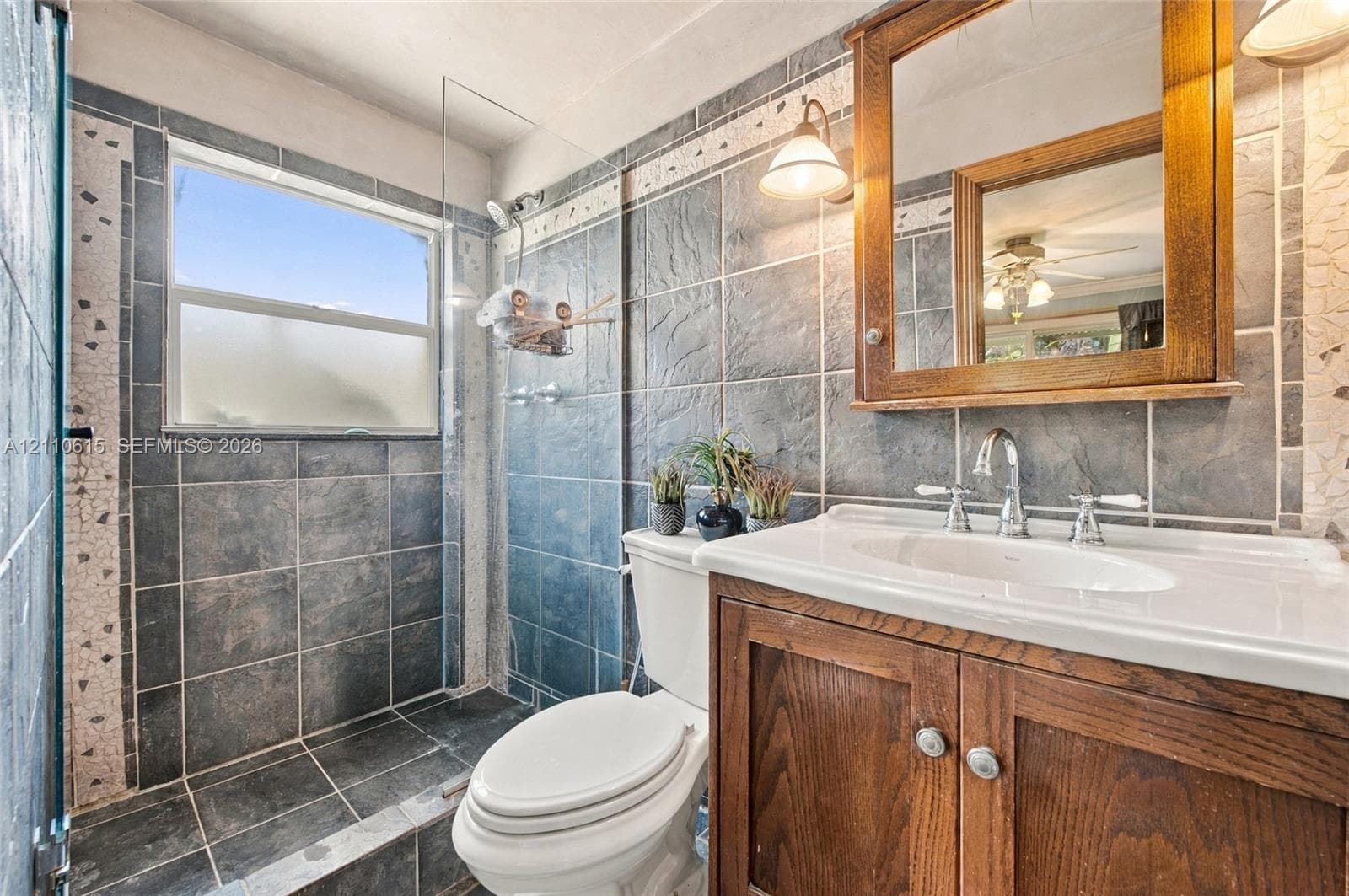 Bathroom featuring large-format dark grey wall and floor tiling, wood-finish cabinetry, a built-in recessed mirror, a white ceramic pedestal-style vanity top, and a glass-enclosed shower