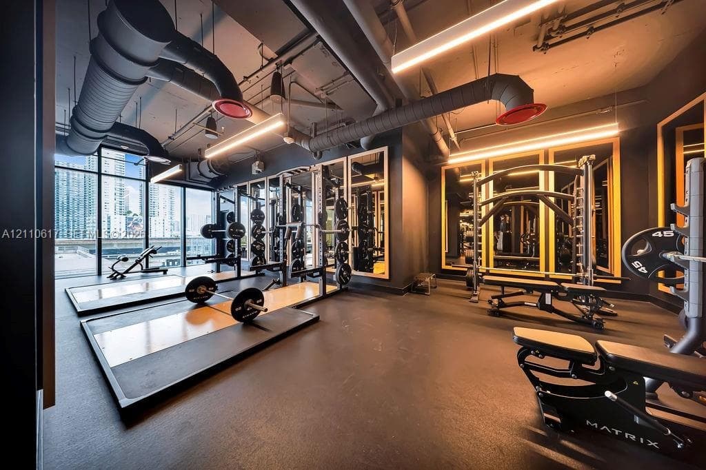Fitness facility featuring rubber flooring, floor-to-ceiling windows, industrial-style exposed ductwork, wall-mounted mirrors, and integrated linear lighting