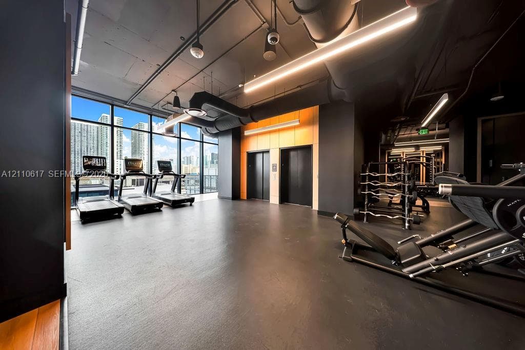Fitness area featuring floor-to-ceiling windows, exposed industrial-style ductwork, high ceilings, rubberized flooring, and dual elevator access