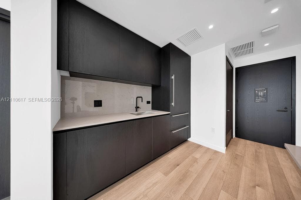 Modern kitchenette featuring matte black flat-panel cabinetry, integrated refrigerator, neutral solid-surface countertop, light wood-finish flooring, and a matte black faucet