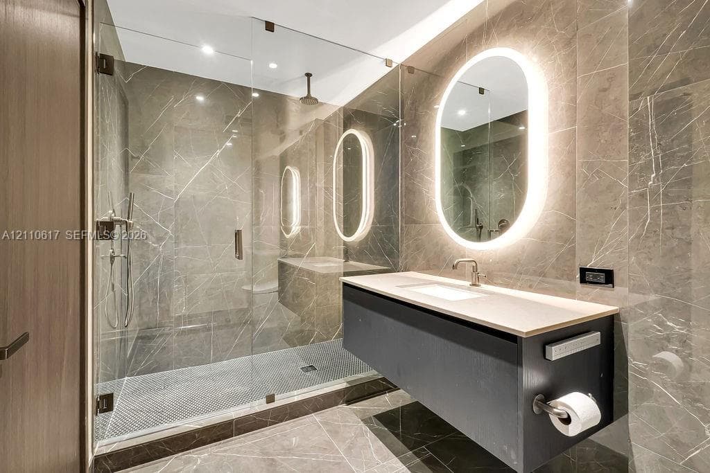 Floating vanity with a light-toned countertop, floor-to-ceiling stone tile wall, walk-in glass-enclosed shower, and oval-shaped backlit mirror