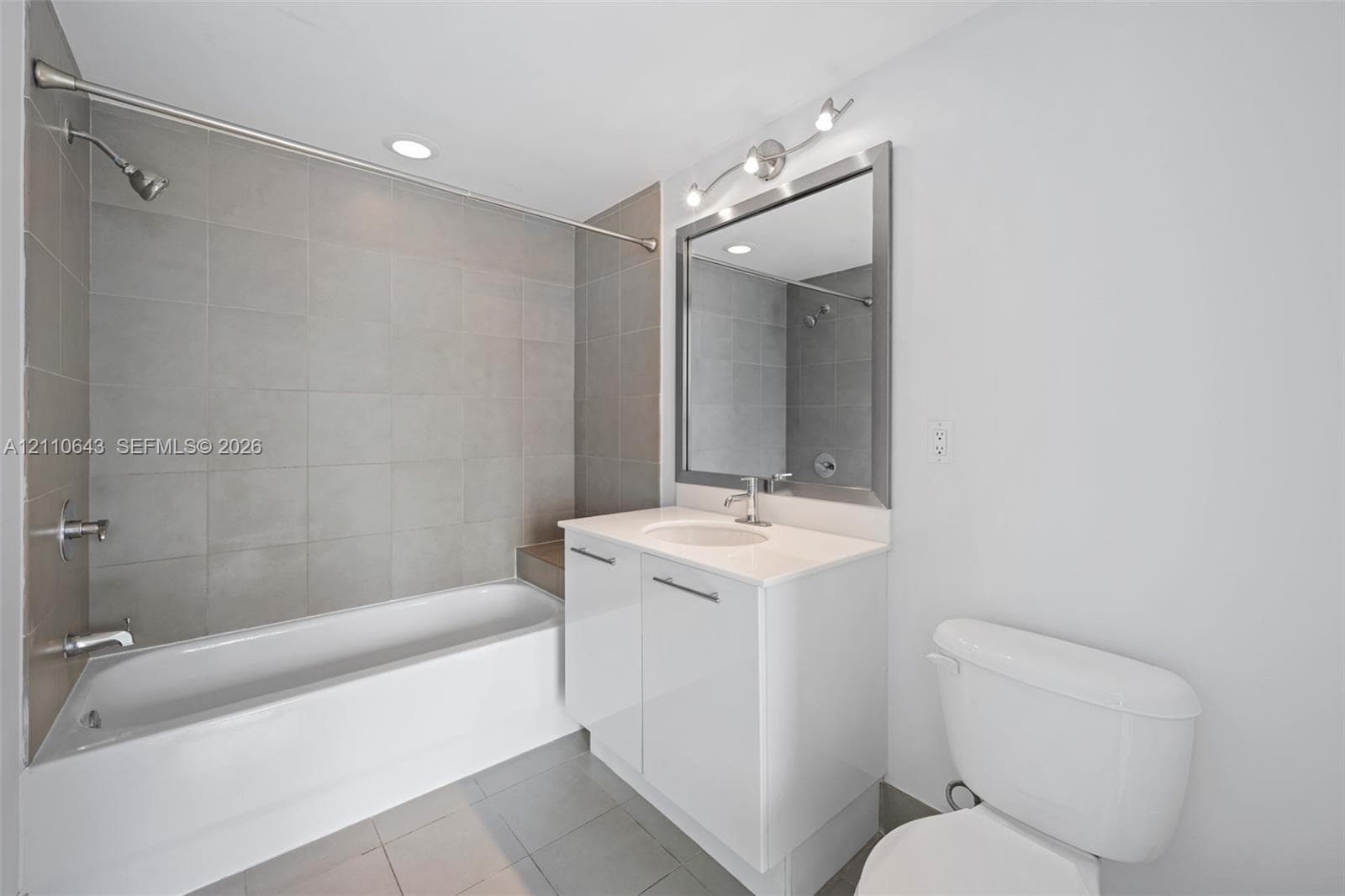 Bathroom featuring a porcelain tile bathtub surround, white vanity cabinet with an integrated sink, polished chrome faucet, framed mirror with integrated lighting, and neutral floor tile