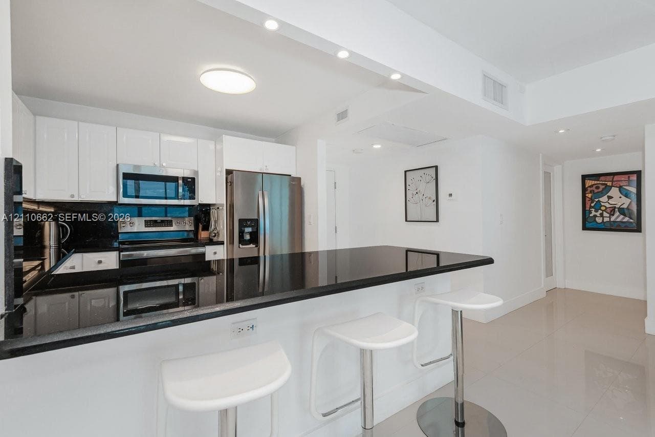 Kitchen featuring white shaker-style cabinetry, dark stone countertops, stainless steel appliances, under-cabinet lighting, and large-format tile flooring