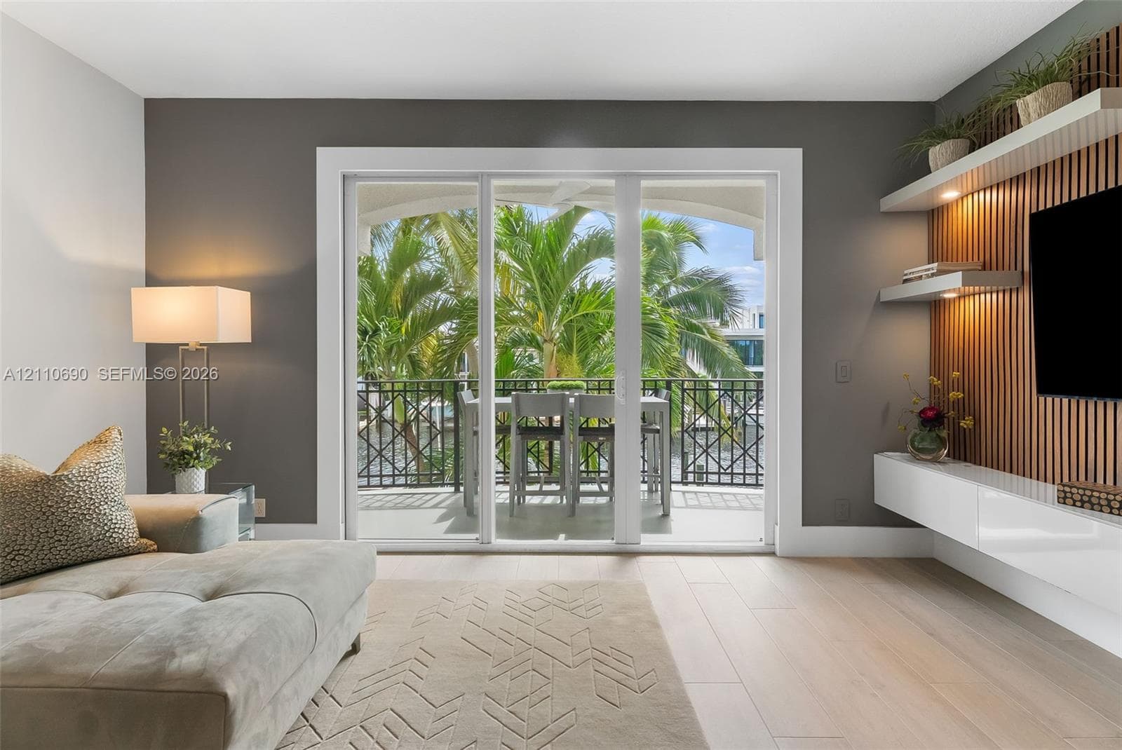 Sliding glass doors opening to a covered balcony with decorative metal railing, wood-slat accent wall with integrated shelving, and white high-gloss floating cabinetry