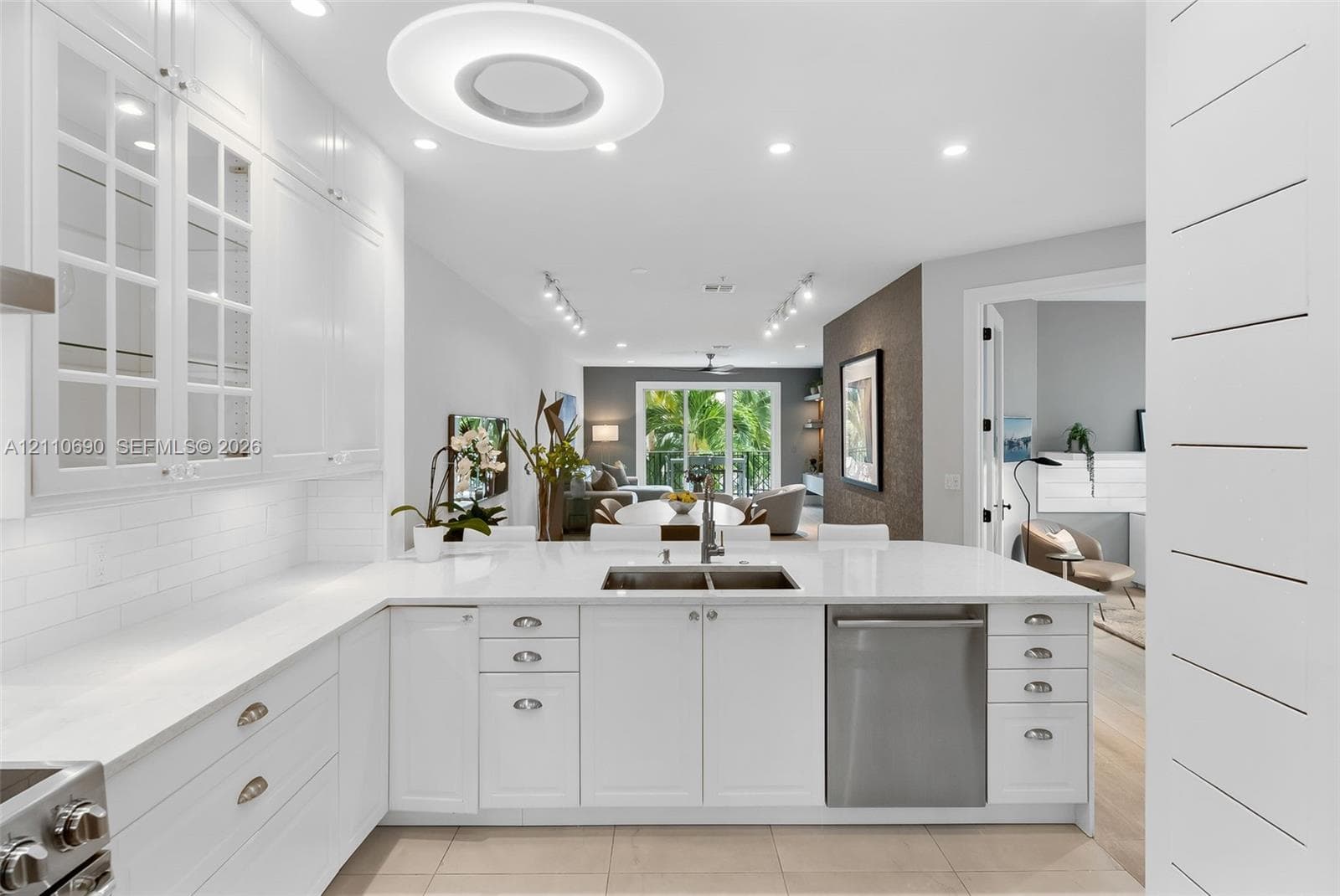 White cabinetry featuring glass-paneled upper doors, expansive countertops with an integrated stainless steel dual-basin sink, stainless steel dishwasher, tile flooring, and decorative horizontal wall paneling
