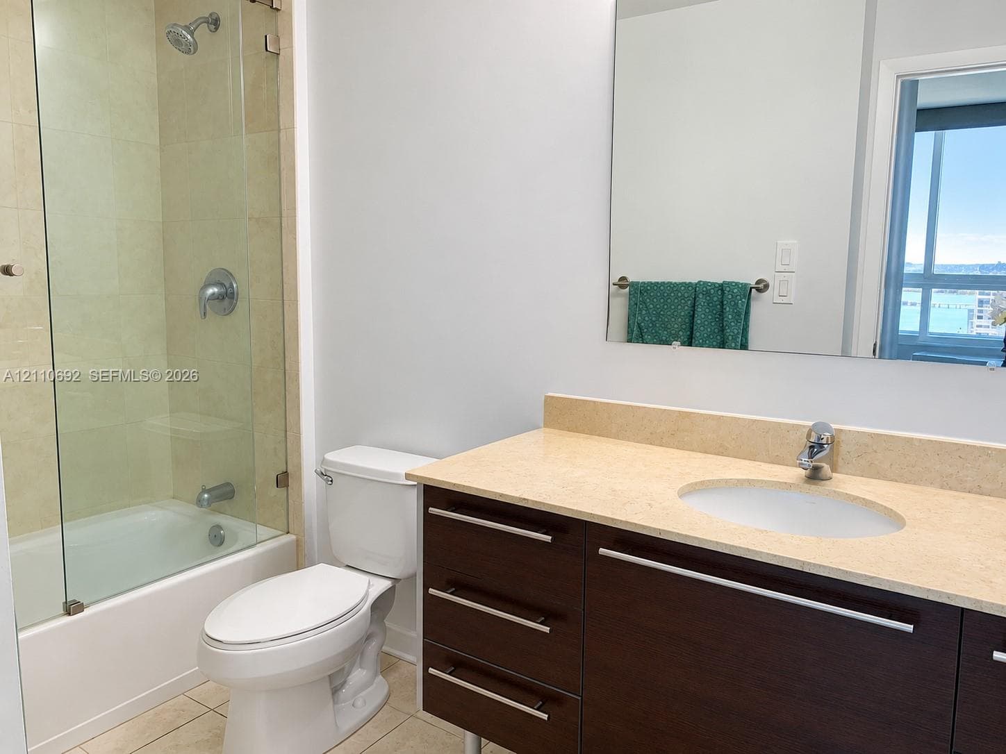 Bathroom featuring a dark wood-finish vanity with a light-toned countertop, integrated oval sink, glass-enclosed shower-tub combination with tile surround, and a wall-mounted mirror