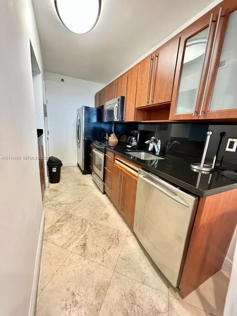 Galley kitchen featuring wood-finish cabinetry, black solid-surface countertops, stone-tile flooring, and stainless steel appliances