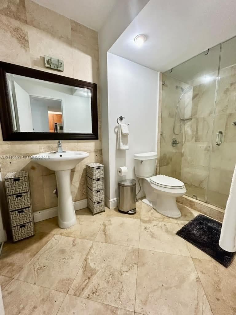 Bathroom featuring natural stone tile flooring and wall accents, a white porcelain pedestal sink, and a glass-enclosed shower with a rainfall showerhead