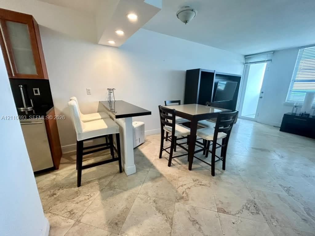 Polished stone flooring, built-in breakfast bar with a dark solid surface countertop, recessed ceiling lighting, and glass-paneled upper cabinetry