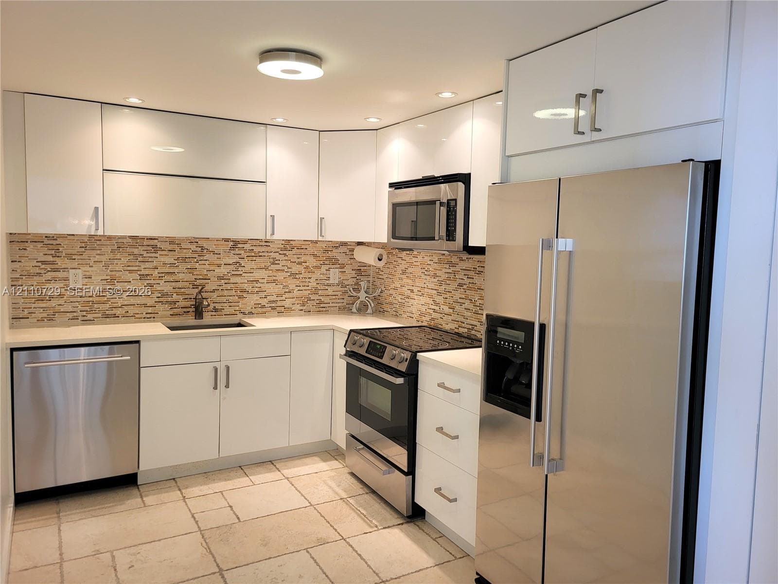 Kitchen featuring high-gloss white cabinetry, stainless steel appliances, stacked stone backsplash, stone-finish flooring, and recessed lighting