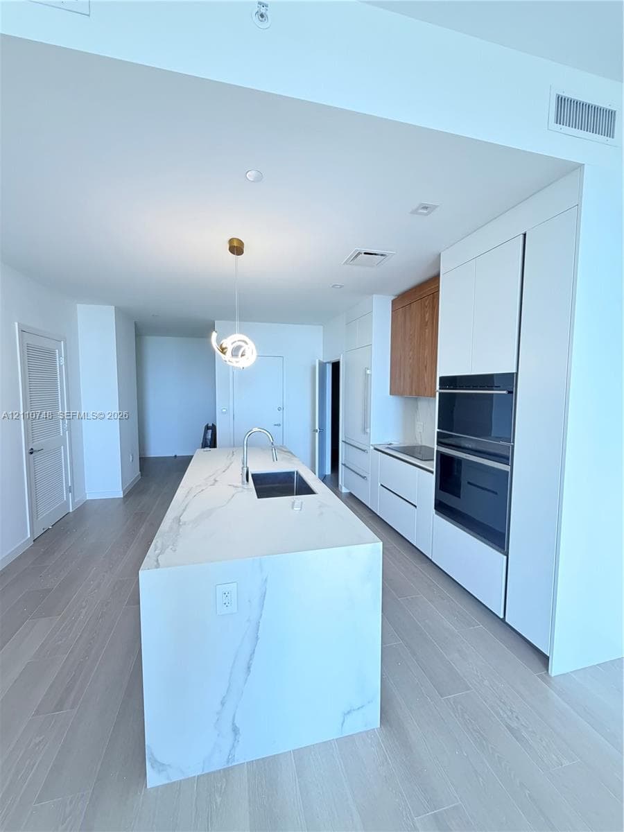 Kitchen featuring a large central island with a waterfall-edge countertop, stone-finish surfacing, integrated sink, and gooseneck faucet, paired with white flat-panel cabinetry, wood-finish accents, and a built-in double oven