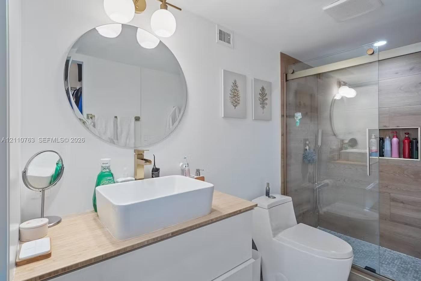 Floating vanity with a light wood-finish countertop, rectangular vessel sink, gold-tone faucet, round wall-mounted mirror, and glass-enclosed shower with wood-finish tile wall and built-in recessed niche
