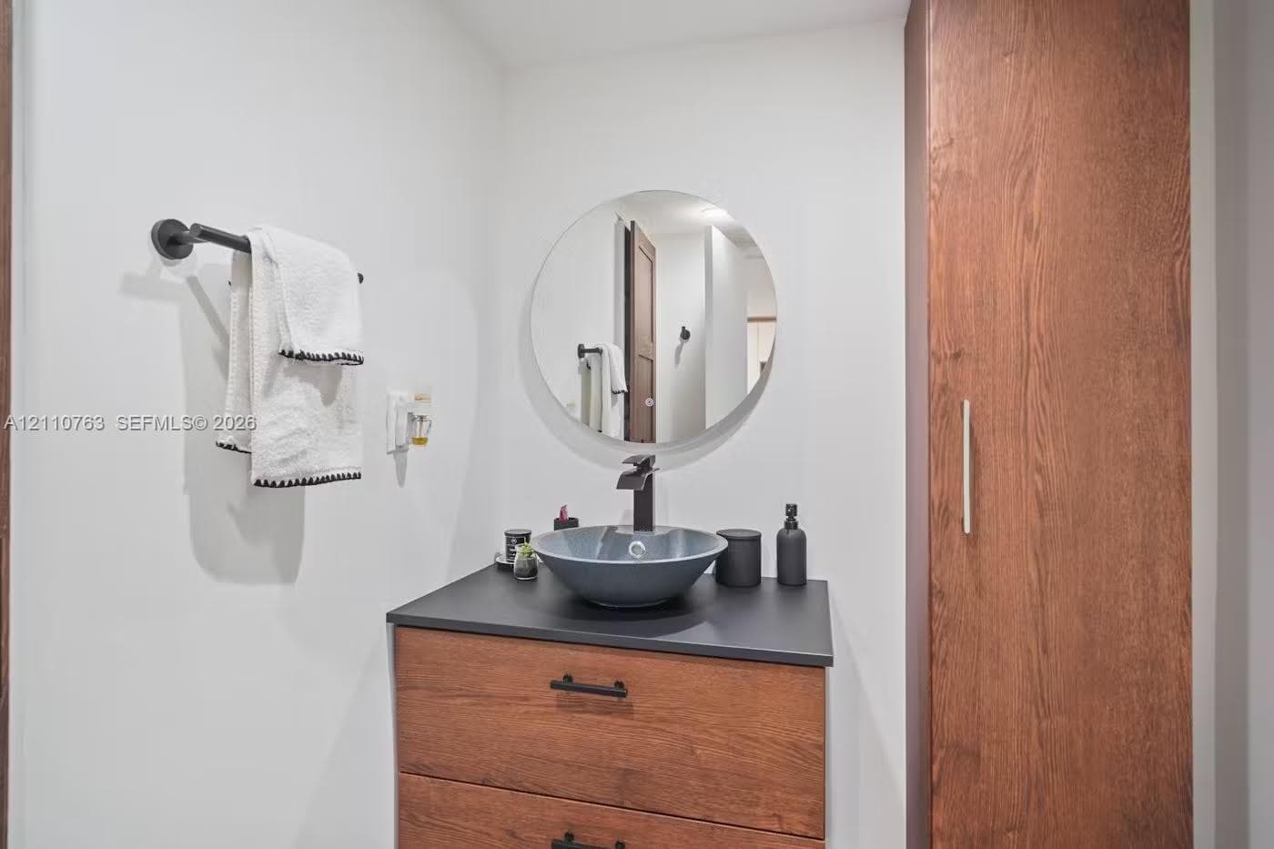 Modern vanity featuring a wood-finish cabinet, dark solid-surface countertop, circular vessel sink, and matte black faucet, complemented by a tall built-in storage cabinet and a circular wall-mounted mirror