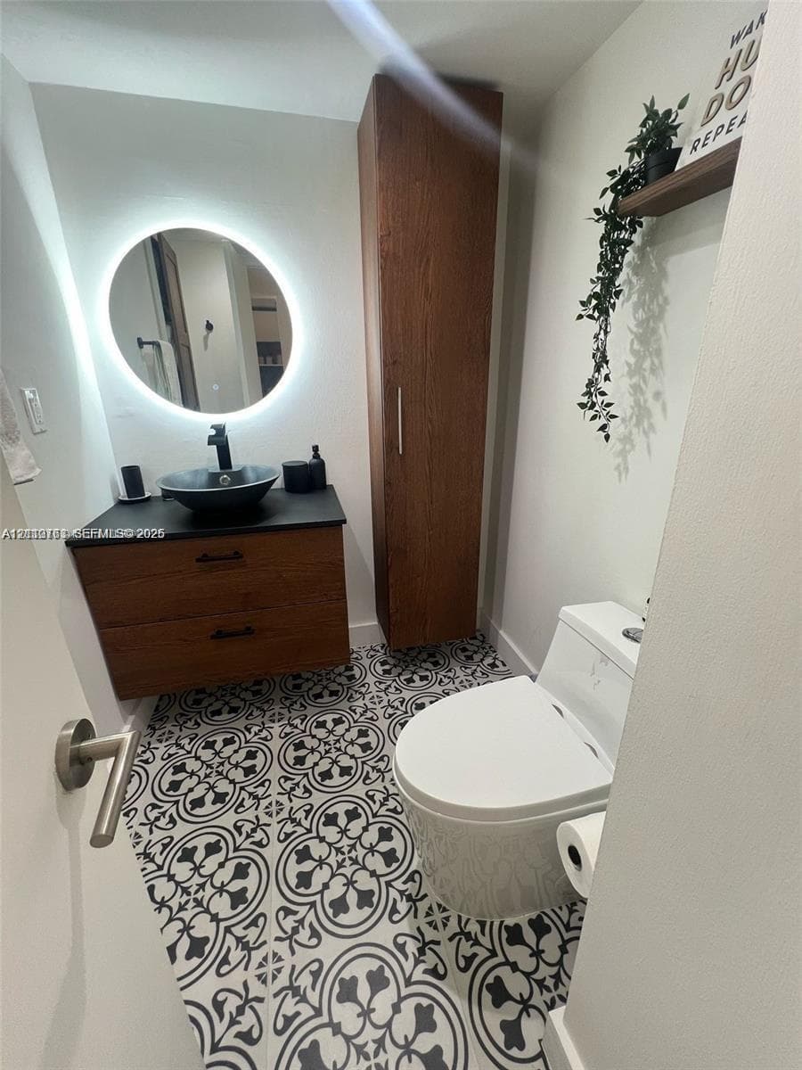 Modern vanity with wood-finish cabinetry, black stone countertop, and circular vessel sink, complemented by a backlit round mirror, floor-to-ceiling wood-finish storage cabinet, and patterned tile flooring