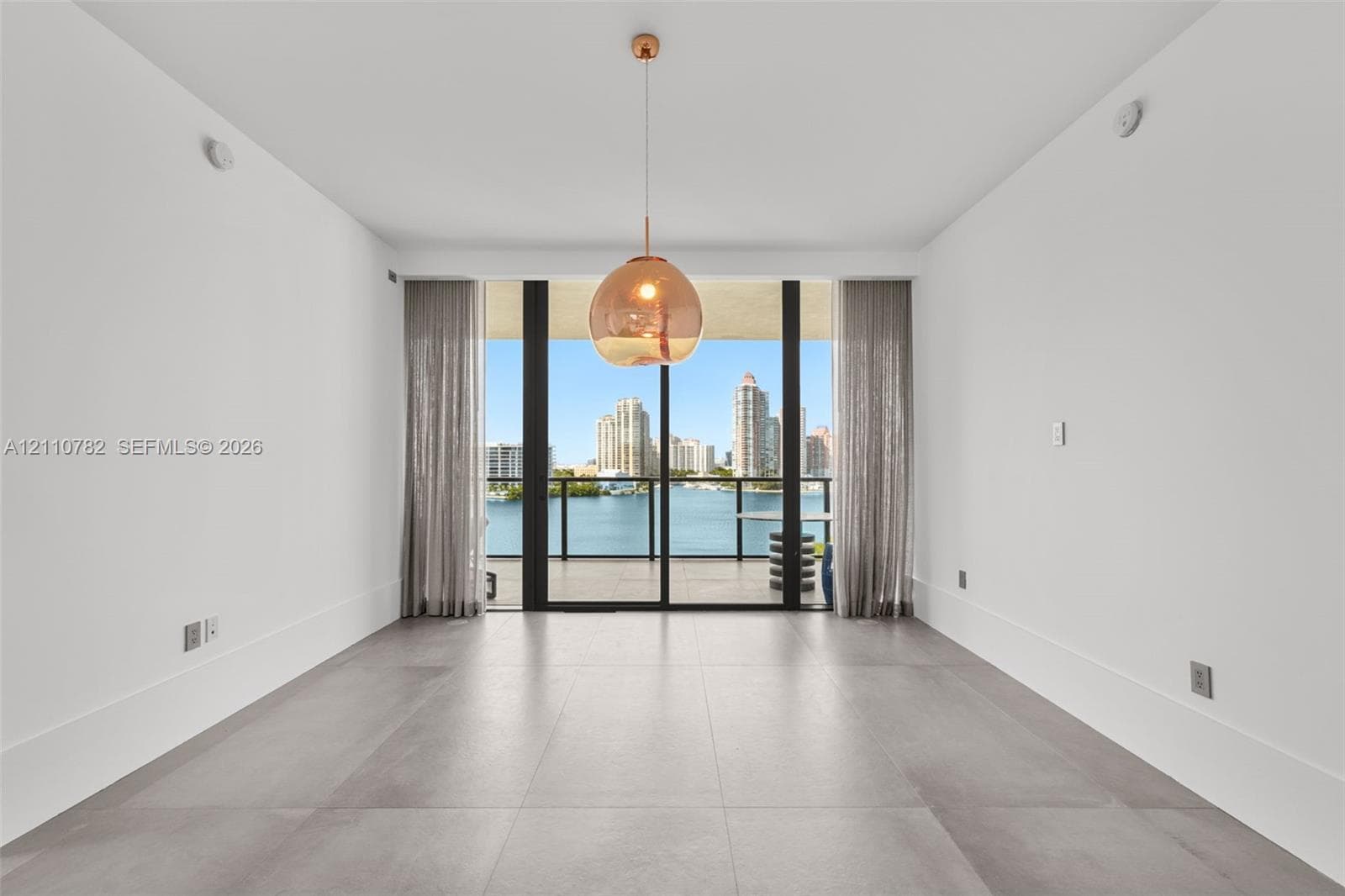 Large-format tile flooring, recessed linear lighting, and expansive floor-to-ceiling glass paneling leading to a balcony with water and skyline scenery