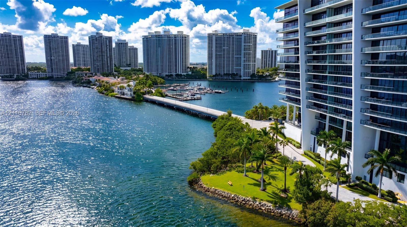 High-rise residential building featuring floor-to-ceiling glass paneling, wrap-around balconies, a porte-cochère entryway, and an outdoor swimming pool, situated on a waterfront lot with dense perimeter foliage