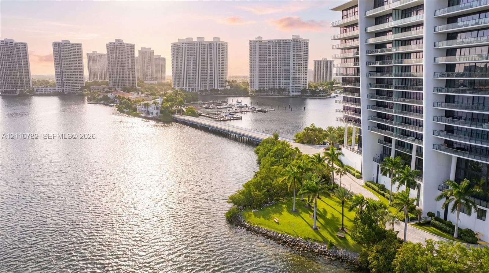 Private waterfront sandy beach featuring a perimeter fence, mature landscaping, and stone bulkhead
