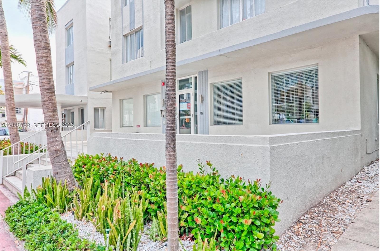 Mid-century modern exterior featuring a smooth stucco facade, multi-pane windows, a recessed glass-paned entrance, and structured tropical landscaping