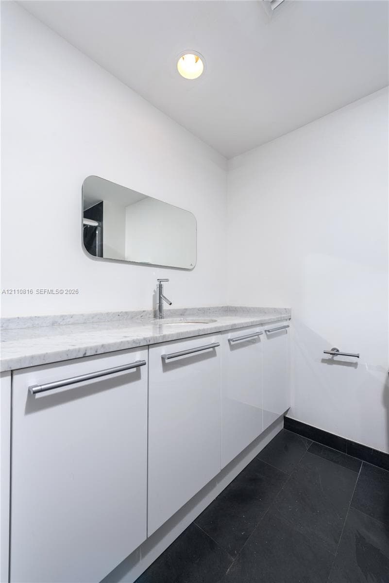 Custom white cabinetry with sleek horizontal pulls, stone-composite countertop, undermount sink, wall-mounted mirror with rounded edges, and dark tile flooring