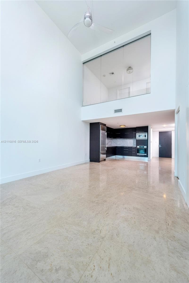 Double-height ceilings featuring an overhead fan, polished stone flooring, dark-toned cabinetry, stainless steel built-in appliances, and a glass-paneled interior wall