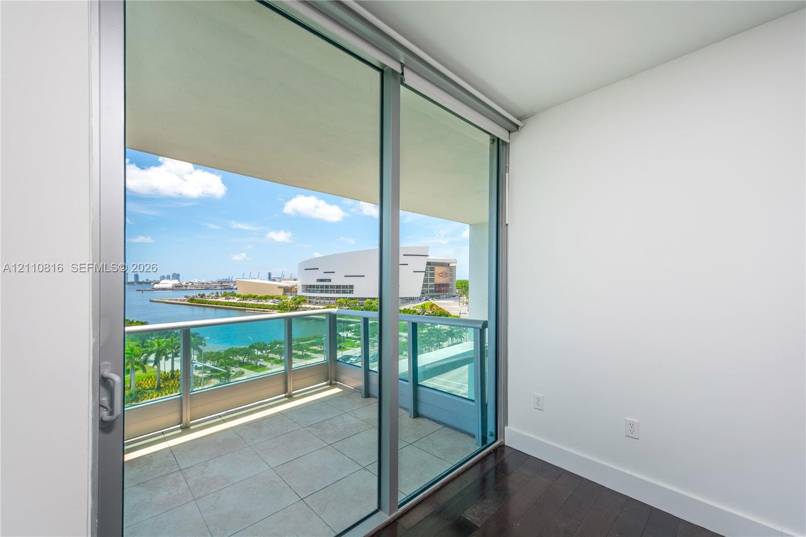Floor-to-ceiling sliding glass door access to a covered balcony with a metal and glass railing, featuring tile flooring and hardwood-finish interior flooring