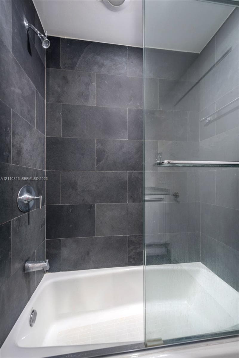 Bathtub with a glass enclosure, featuring large-format dark stone wall tile, chrome shower head, and wall-mounted chrome hardware