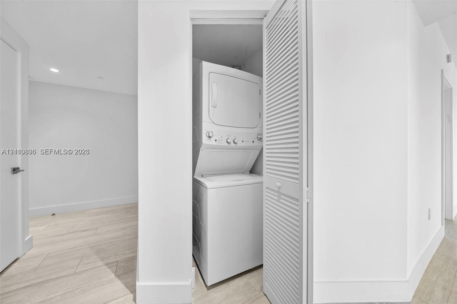 Laundry closet featuring a white stacked washer and dryer unit enclosed by bi-fold louvered doors, situated over light-toned wood-finish flooring