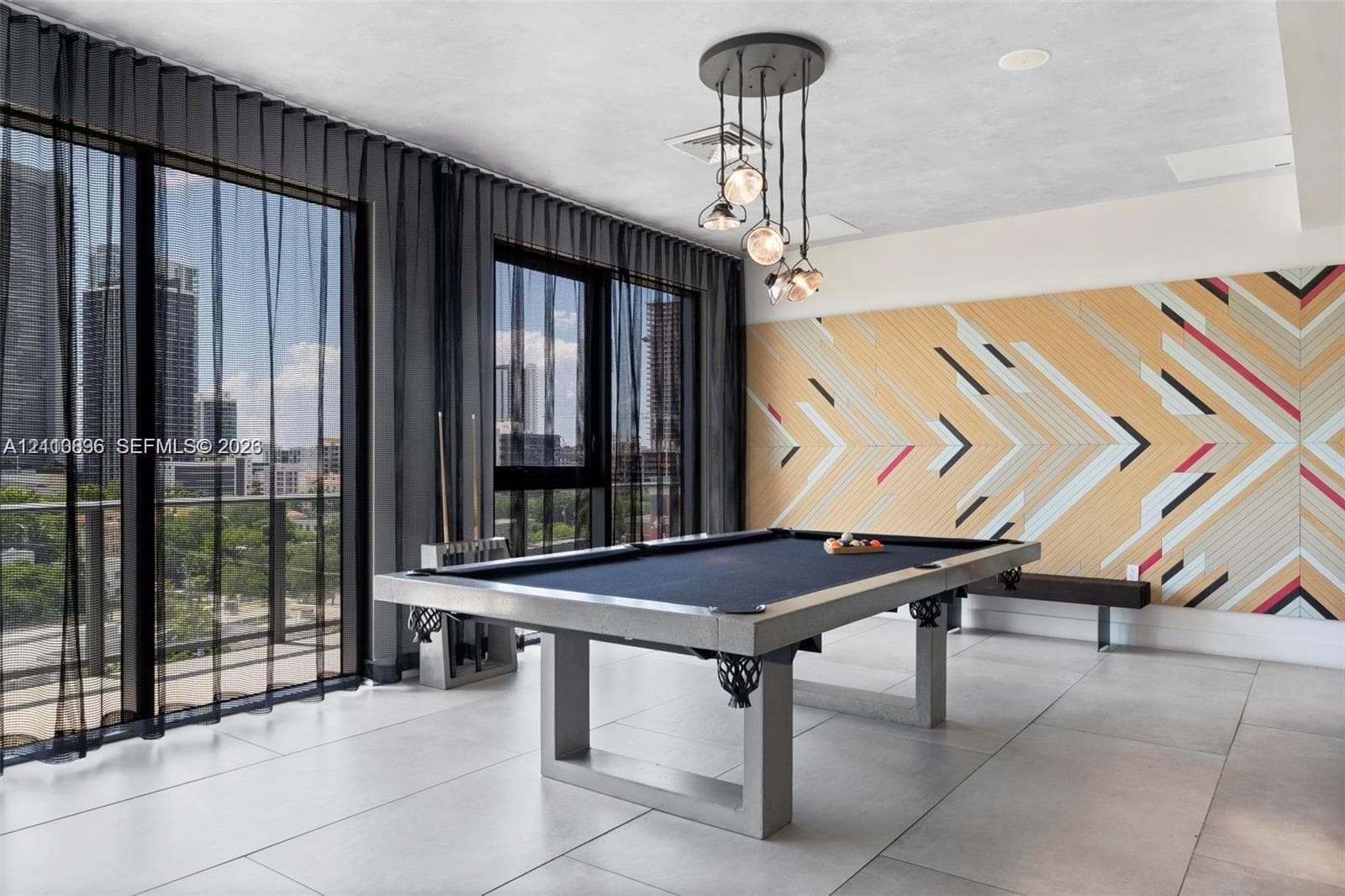Modern recreation room featuring a regulation pool table, geometric wood-finish accent wall with multi-colored chevron detailing, floor-to-ceiling glass doors with dark framing, polished concrete flooring, and a contemporary pendant light fixture suspended from the ceiling