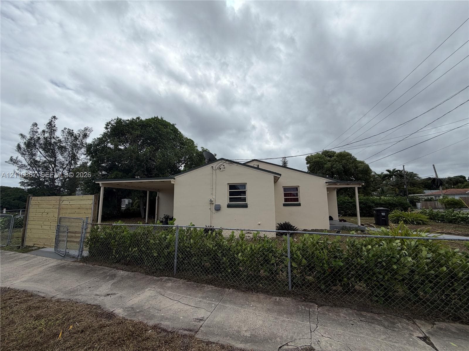 Stucco exterior featuring a covered carport, wood privacy fencing, chain-link perimeter fencing, and mature trees