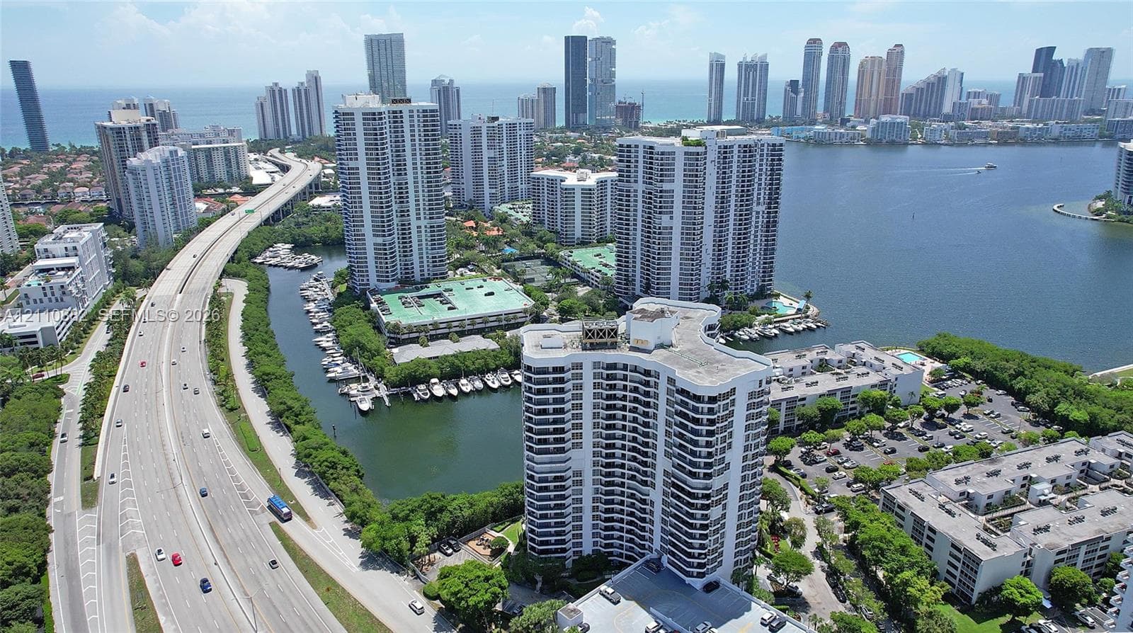 Aerial perspective of high-rise building with balcony tiers, situated along a coastal waterway with surrounding urban infrastructure and tropical landscaping