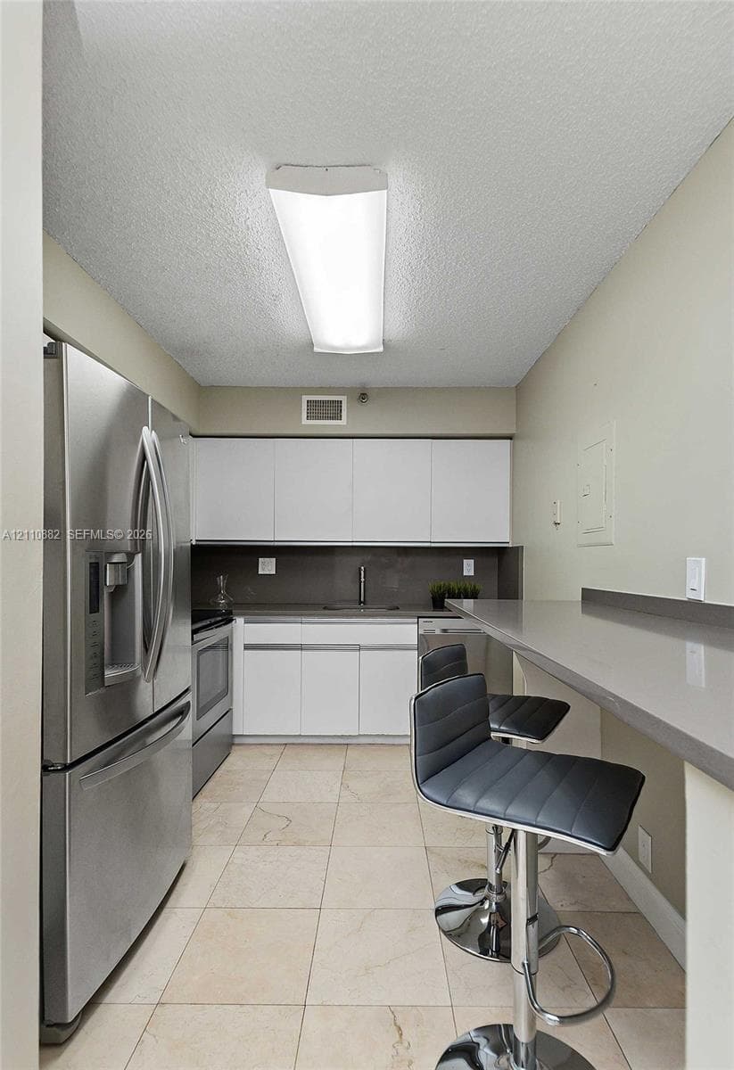 Kitchen featuring white flat-panel cabinetry, stainless steel refrigerator, solid-surface countertop with integrated breakfast bar, and tile flooring