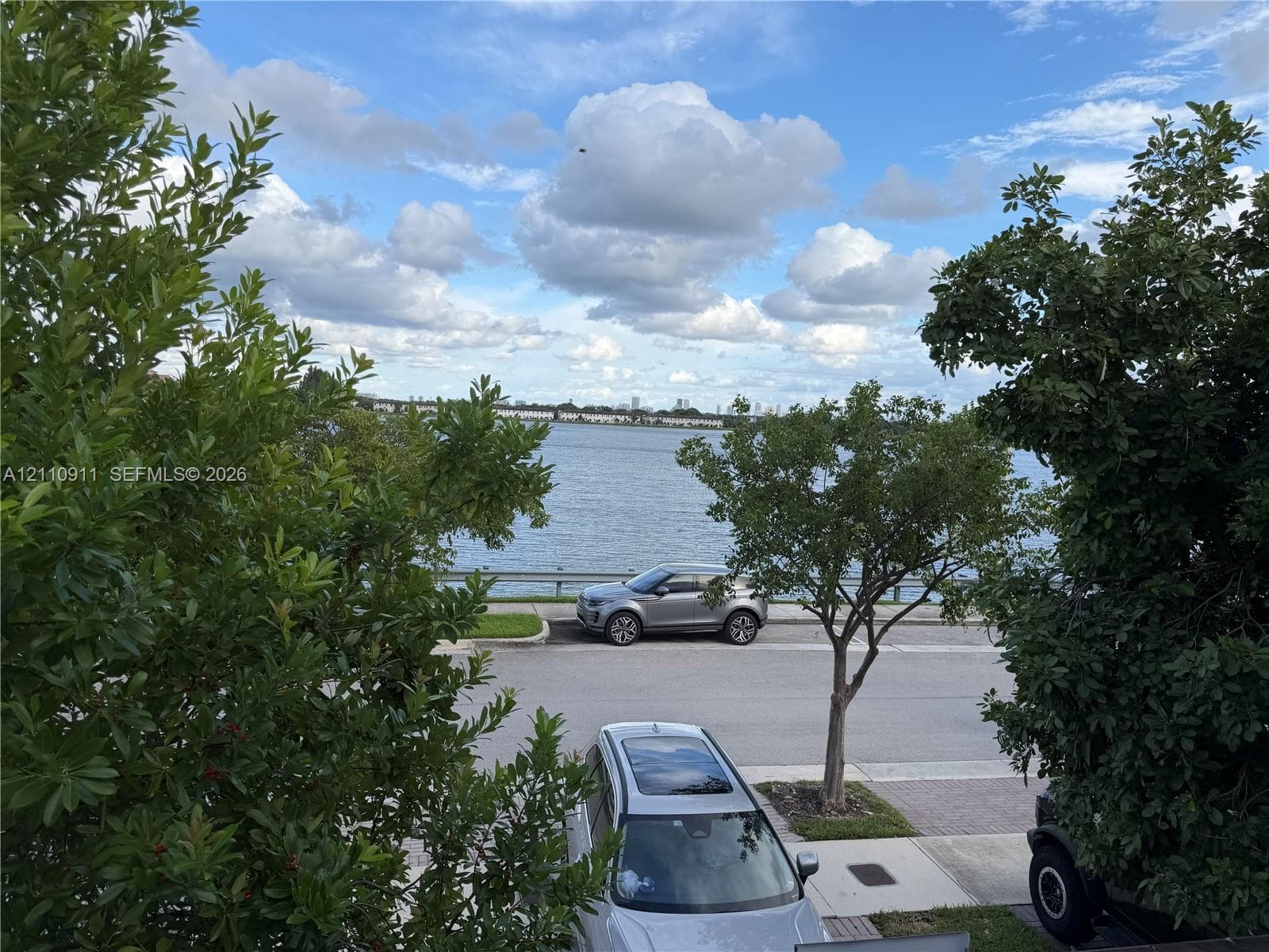Waterfront location featuring a paved roadway, concrete curbing, mature trees, and metal guardrail