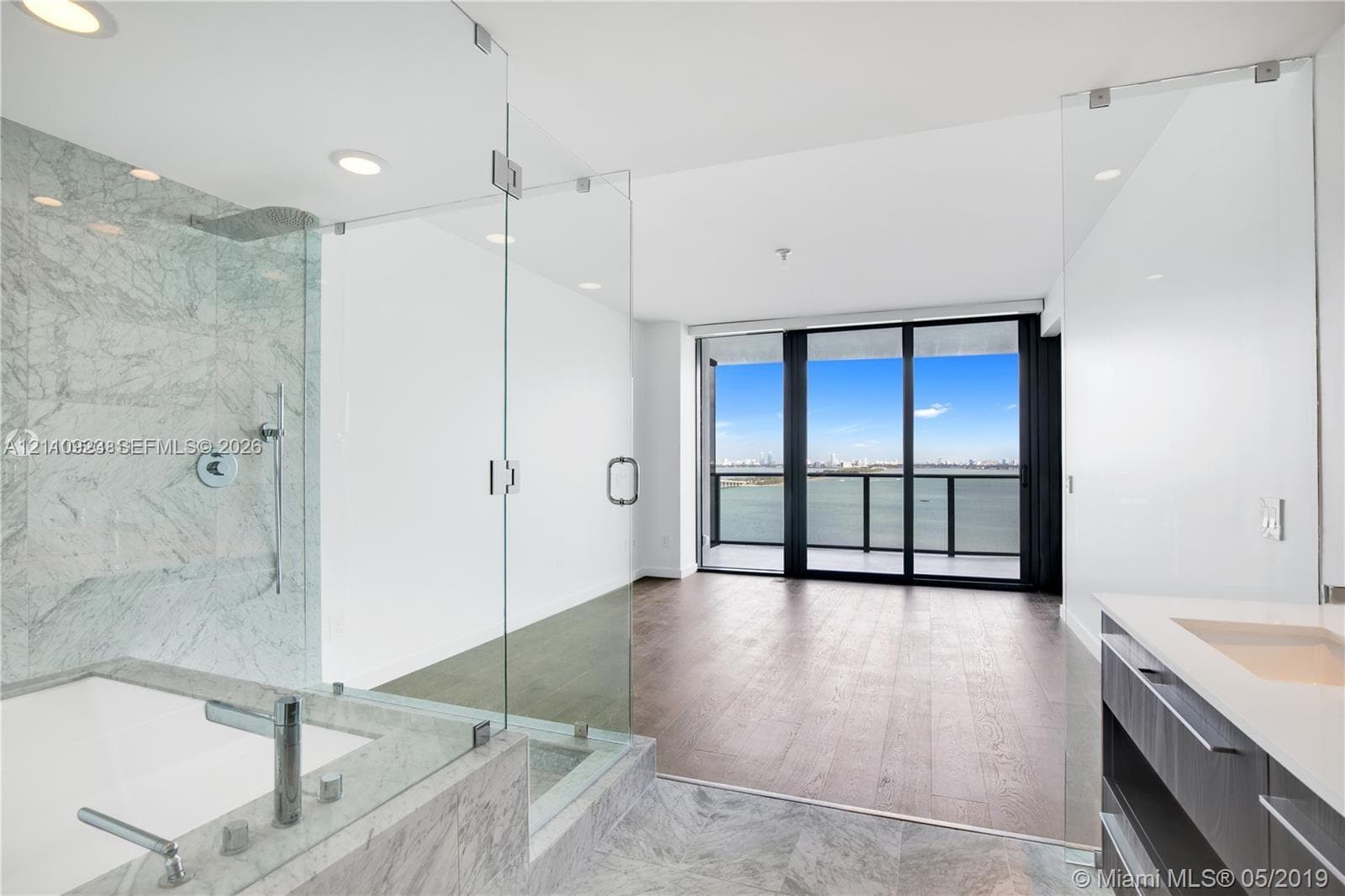 Bathroom featuring stone-surround soaking tub, glass-enclosed walk-in shower with stone wall detailing, wood-finish flooring, and floor-to-ceiling windows with water backdrop