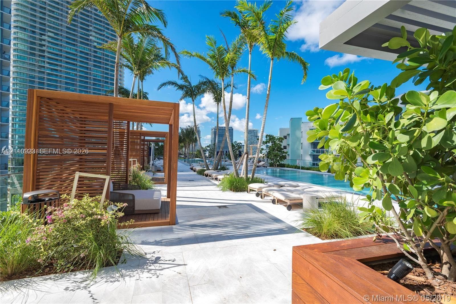 Outdoor swimming pool featuring a stone-tiled deck, wood-slatted cabanas, integrated wood-finish planters, and mature palm trees