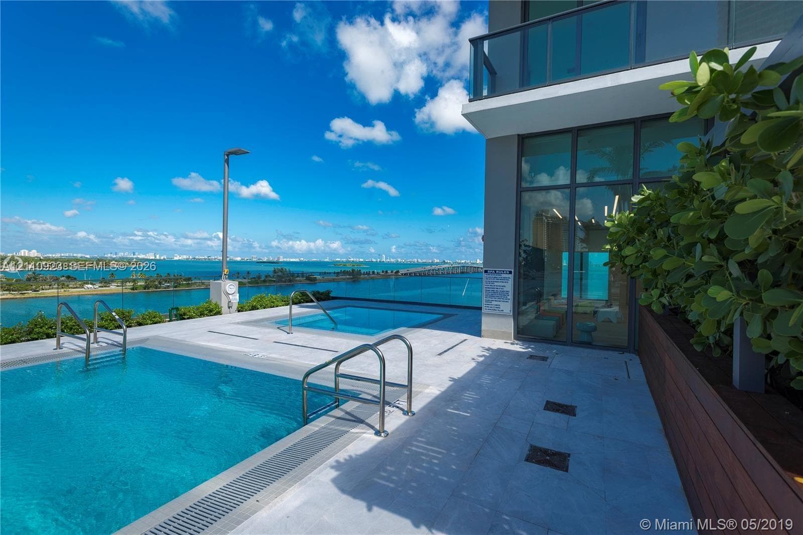 Terrace level featuring an infinity-edge swimming pool, elevated spa, light-toned stone decking, glass panel railings, and metal-framed floor-to-ceiling windows