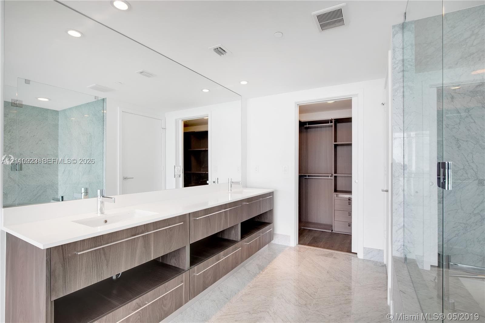 Double vanity with an expansive wall-to-wall mirror, under-mounted sinks, stone-surface countertop, dark wood-finish cabinetry, and glass-enclosed shower with tiled wall surrounds