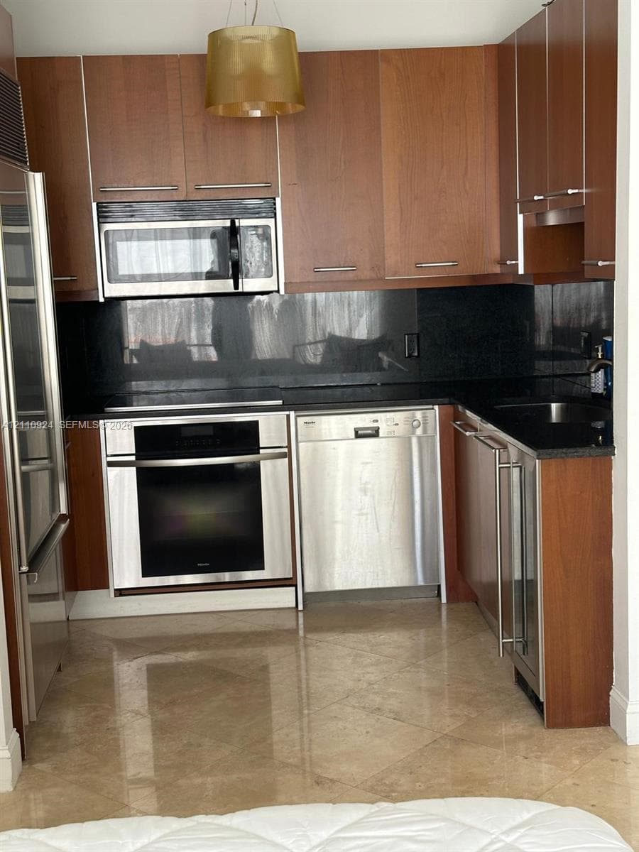 Kitchen featuring wood-veneer cabinetry, stainless steel appliances, dark stone countertops, and polished stone flooring