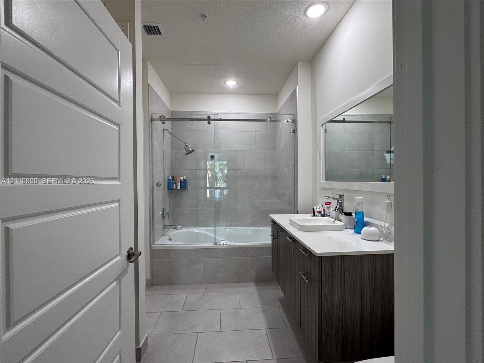 Bathroom featuring a vanity with wood-finish cabinetry, white countertop, rectangular sink, full-width mirror, and a tub-shower combination with a sliding glass door and large-format tile surround