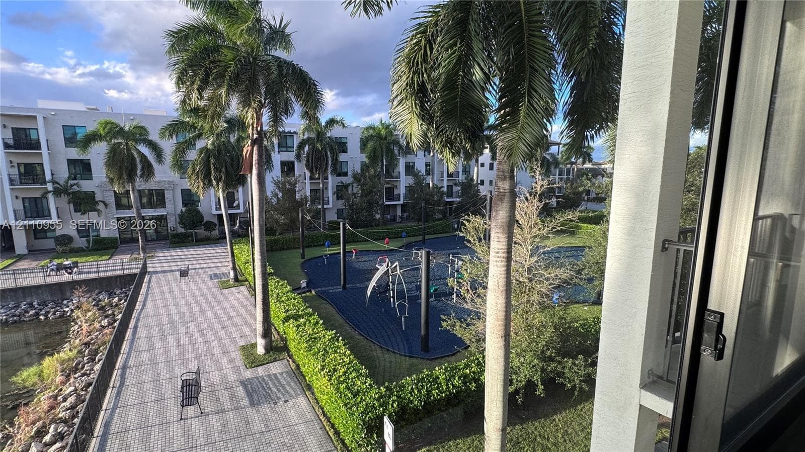 Exterior space featuring a paved walkway, mature palm trees, manicured hedges, and a recreation area with a rubberized surface