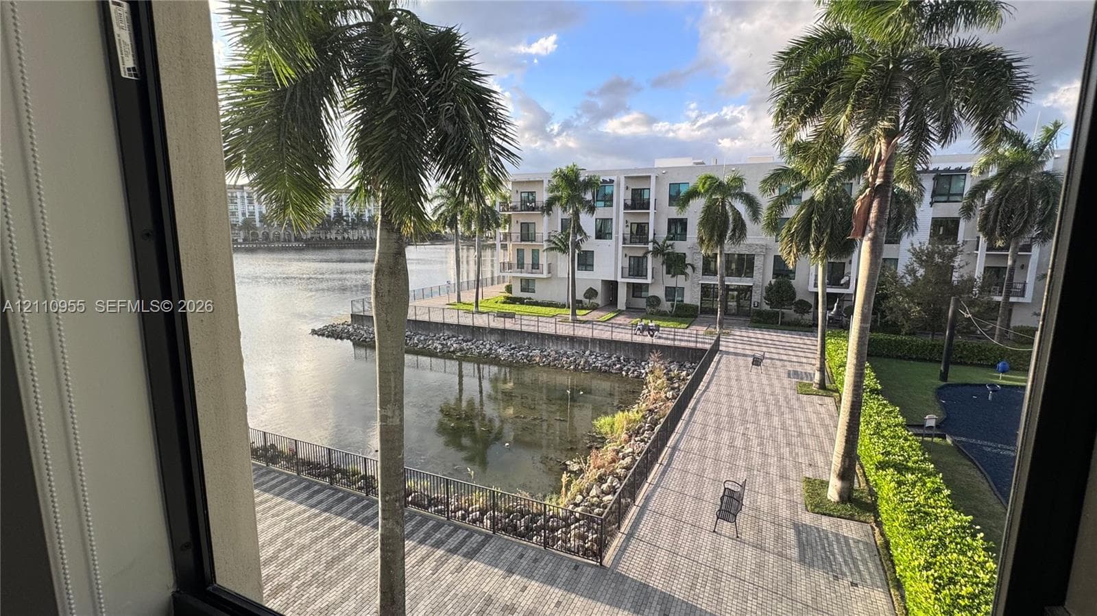 Waterfront property featuring a stone-paver walkway, metal perimeter fencing, mature palm trees, and manicured landscaping